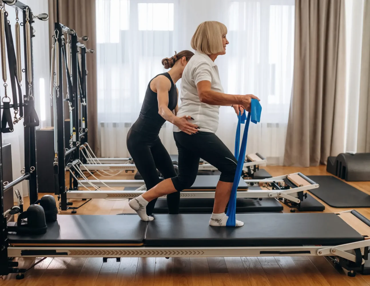Blue colored resistance loops in hands for the legs. Female trainer is helping old woman to exercise.